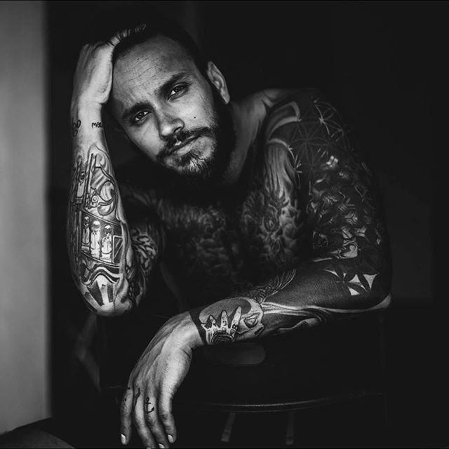 A tattooed man poses for a black and white portrait in a studio setting.
