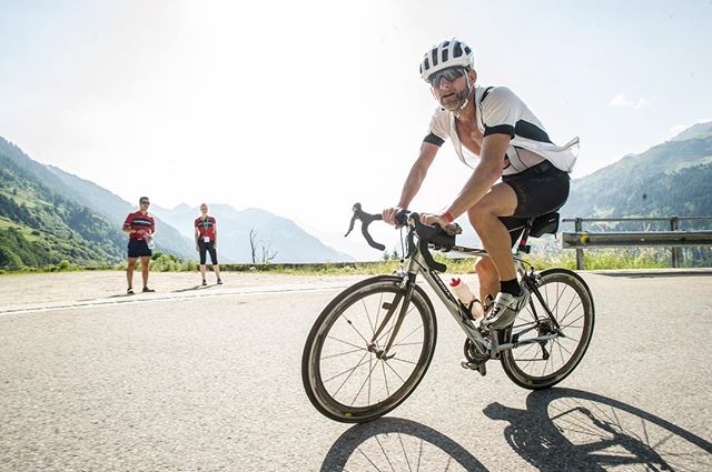A determined cyclist pedals uphill on a scenic mountain road, with two people standing in the background.