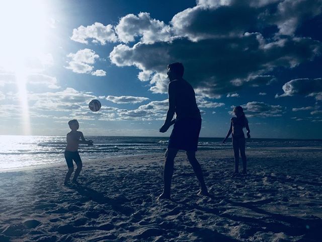 A family is playing soccer on a sunny beach with a blue sky and fluffy clouds.