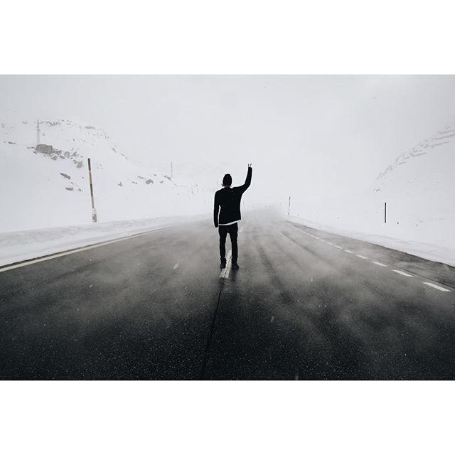 A man stands on a snowy mountain road, hand raised, in a serene winter landscape.