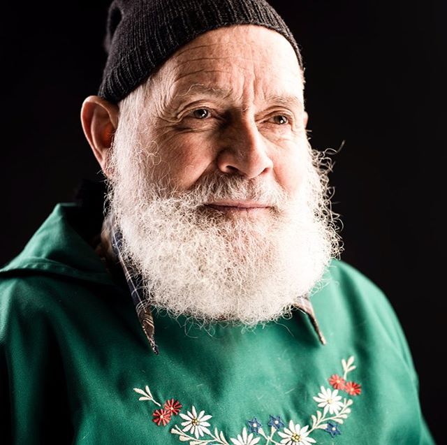 Portrait of an elderly man with a white beard wearing a beanie and an embroidered green top.