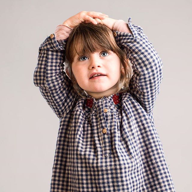 A cute little girl with blue eyes wearing a checked dress poses in a bright studio setting.