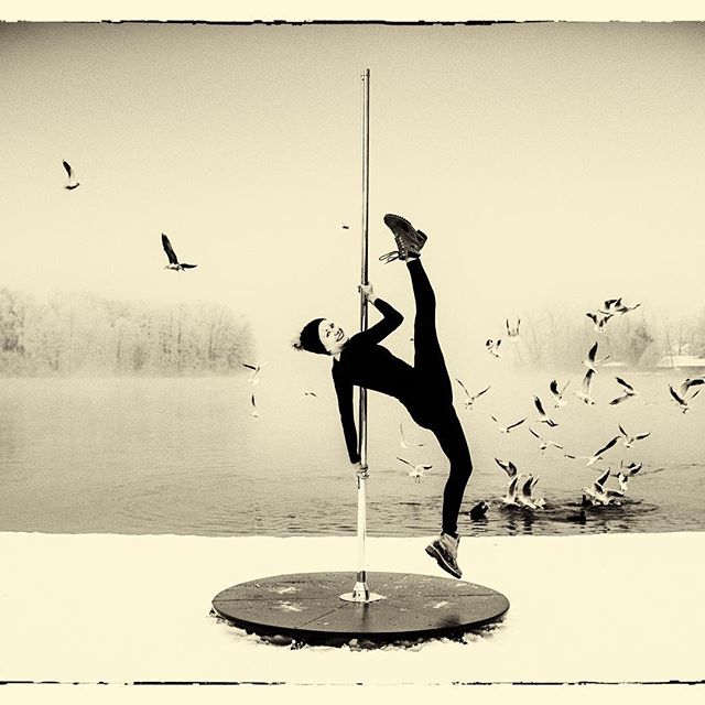 A woman pole dances in a snowy outdoor setting near a lake, surrounded by birds in a vintage black and white style.
