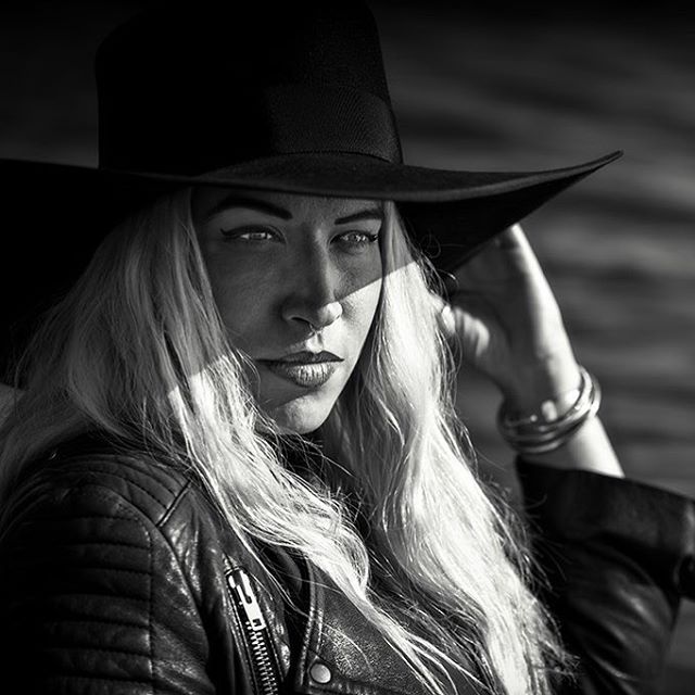 A woman in a black hat and leather jacket poses in a black and white editorial style photograph.