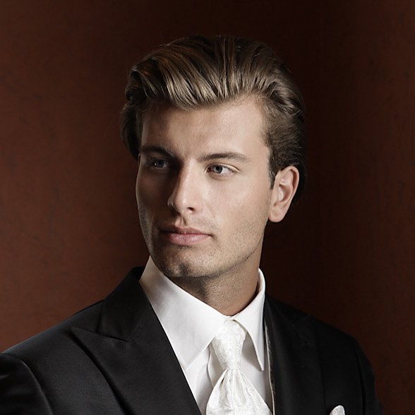 Elegant man in a tuxedo with a white tie poses against a brown background in a studio.