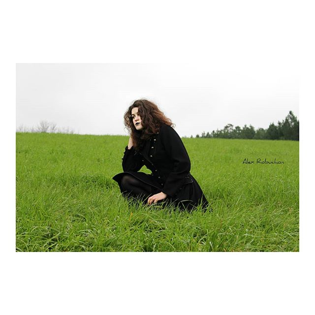 A woman with curly hair in a black dress sits in a green field under a cloudy sky.