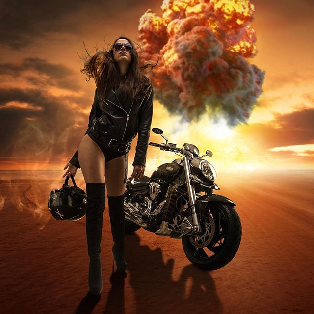 A woman stands by her motorcycle as an explosion erupts in the desert. She is wearing sunglasses, a jacket, and boots.