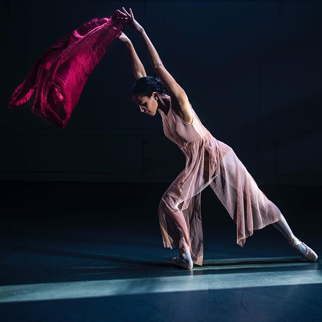 A ballerina poses gracefully with a red scarf in a dance studio, lit with side lighting.