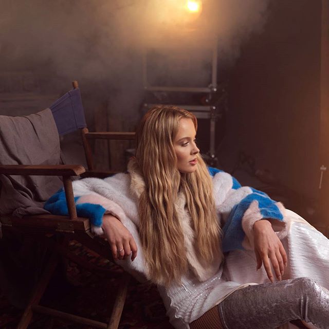 A blonde woman poses elegantly in a fur coat for an editorial fashion shot in a studio setting.