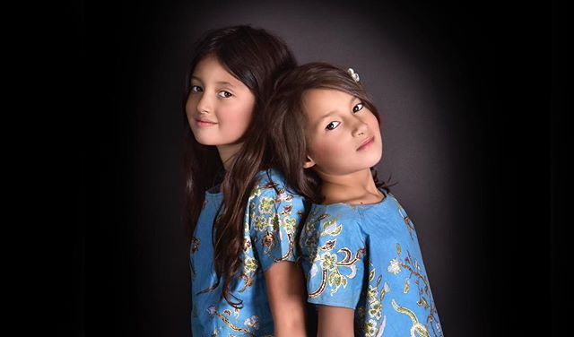 Two young girls stand back to back in a studio setting with a black background, wearing matching blue dresses.