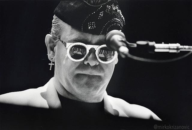 Black and white portrait of Elton John wearing glasses and a hat on stage during a concert performance.