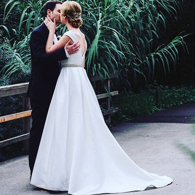 A bride and groom share a kiss on their wedding day, surrounded by lush greenery.