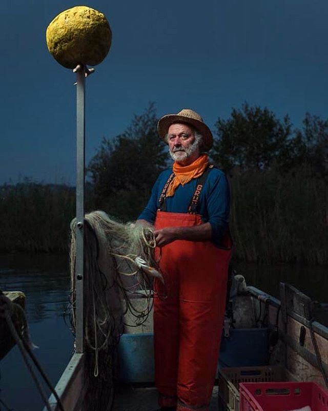 A fisherman in overalls stands on his boat at night, holding his fishing net.