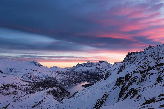 Panoramic view of a snow-covered mountain range with a tranquil lake under a colorful sky at dusk, creating a serene landscape.