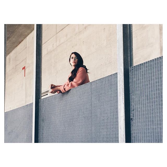 A woman with glasses and a pink jacket on a balcony looks thoughtfully towards the light.