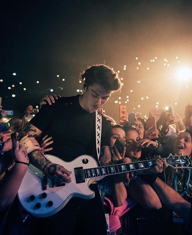A musician plays guitar onstage surrounded by fans at a lively concert venue.