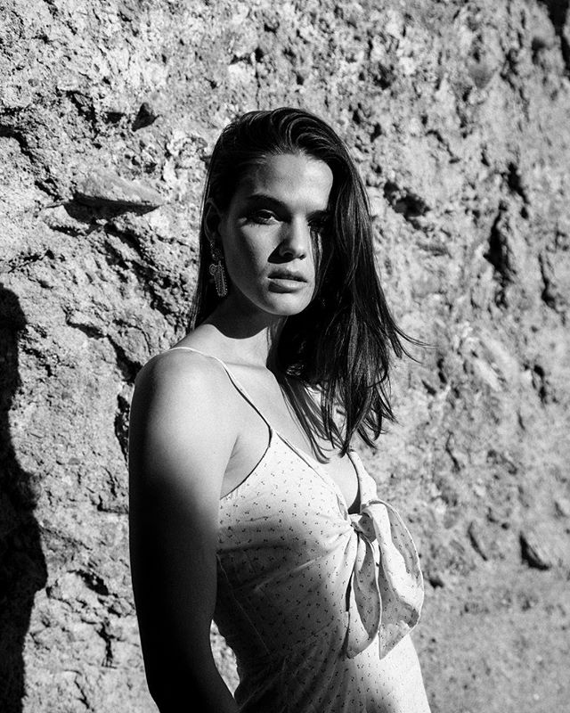A brunette woman poses in a polka dot dress against a textured stone wall in a black and white editorial style.