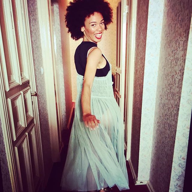 A smiling woman with curly hair poses in a hallway wearing a dress.