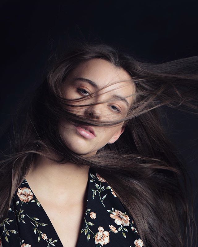A brunette woman with windblown hair poses in a floral top against a dark background.