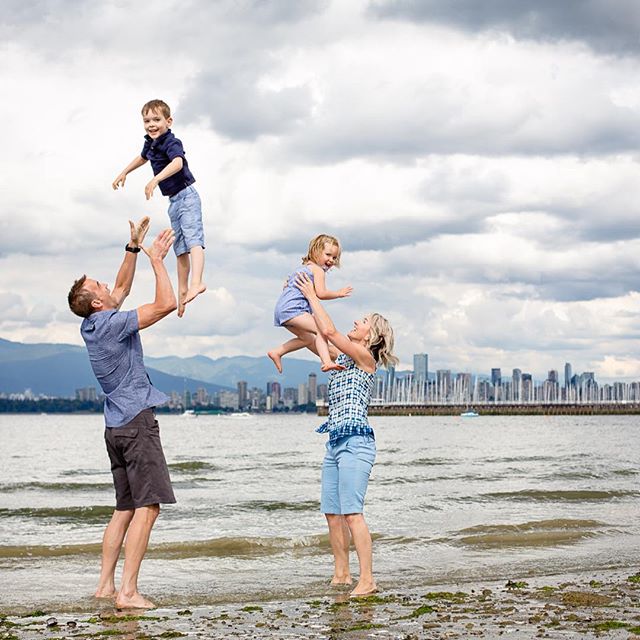 A family of four plays together on a beach, with parents throwing children in the air against a cityscape backdrop.