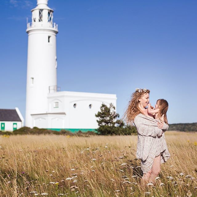 A woman holds a child in a field with a lighthouse in the background on a sunny day.