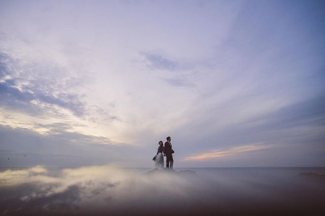 A bride and groom stand together on a misty landscape against a serene sky at sunset.