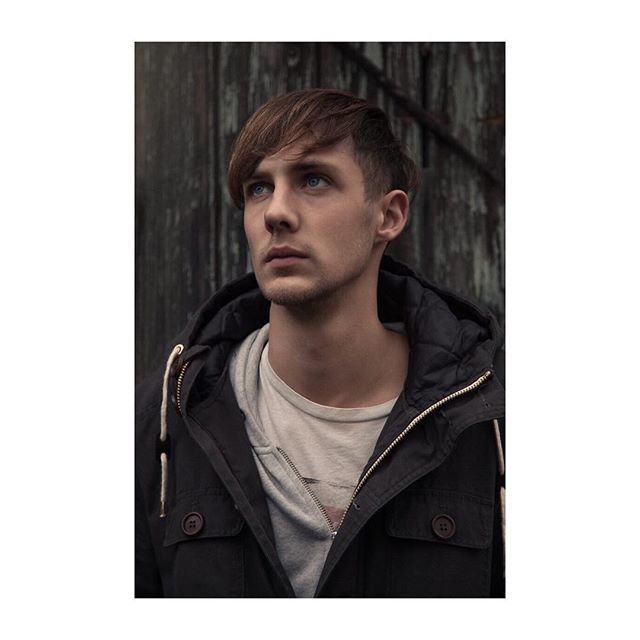 Pensive young man in jacket stands outdoors, looking away with a thoughtful expression.
