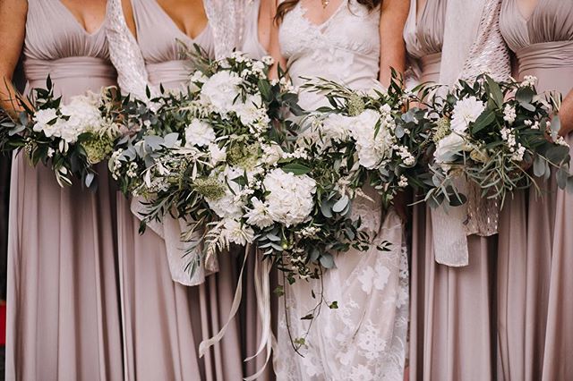 Bridesmaids in taupe dresses hold white floral bouquets at a wedding.