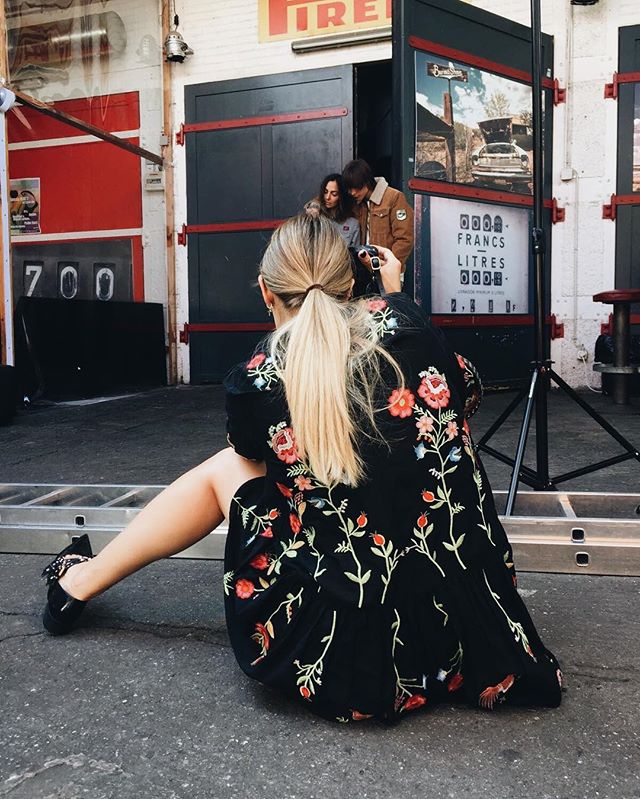 A woman in a floral dress photographs a couple in front of a vintage-style gas station.