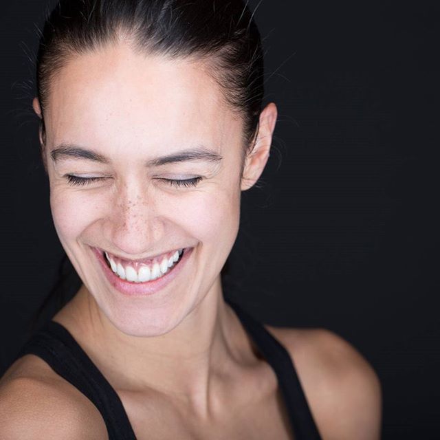 A smiling woman with closed eyes poses in a studio setting against a black background.
