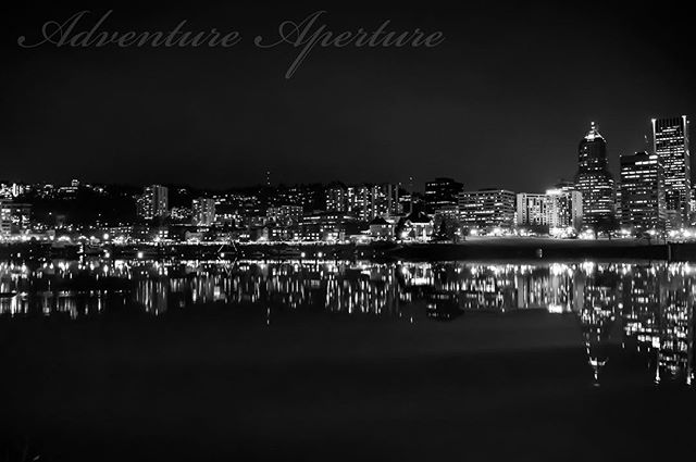 A nighttime cityscape featuring a skyline reflected in the water below in black and white, creating a serene urban scene.