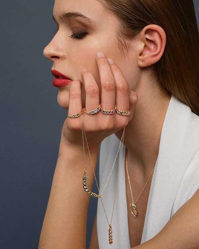 A woman showcases elegant gold rings and necklaces with colorful gemstones in a studio setting.