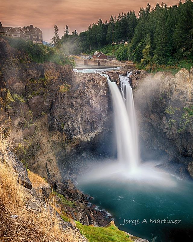 A scenic long exposure captures a powerful waterfall cascading into a pool surrounded by cliffs and forest.