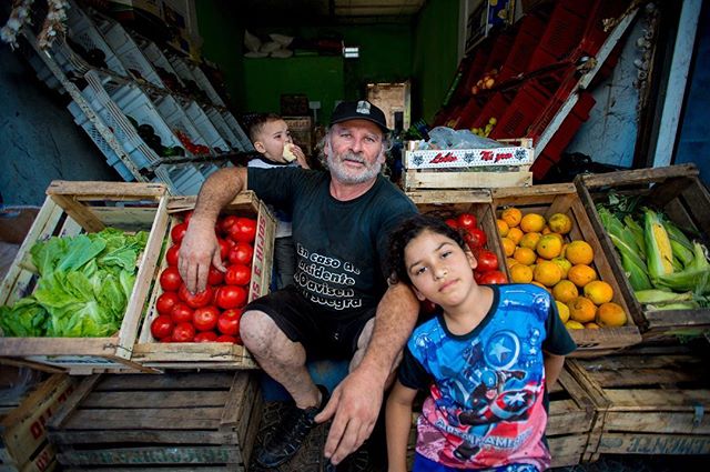 A small business owner poses with two children in front of a fresh produce display at an outdoor market.