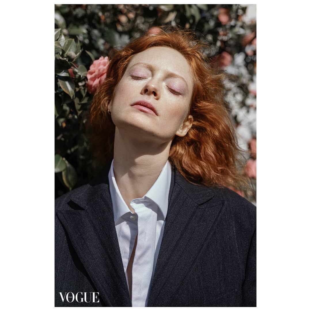A redhead woman in a suit poses serenely with eyes closed in front of a blooming bush for a Vogue fashion editorial.