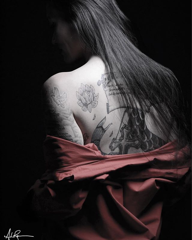 A woman with intricate tattoos poses with a red garment against a dark backdrop, highlighting the art on her back.