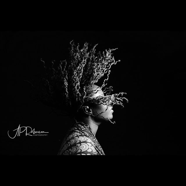 Monochromatic portrait of a woman with dreadlocks in a striking artistic pose against a dark background.