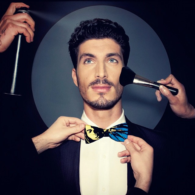 A stylish man is being groomed with a bowtie, makeup, and hairspray in a studio setting.