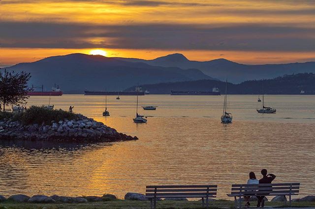 A couple sits on a bench enjoying a beautiful sunset over the ocean with boats and mountains.