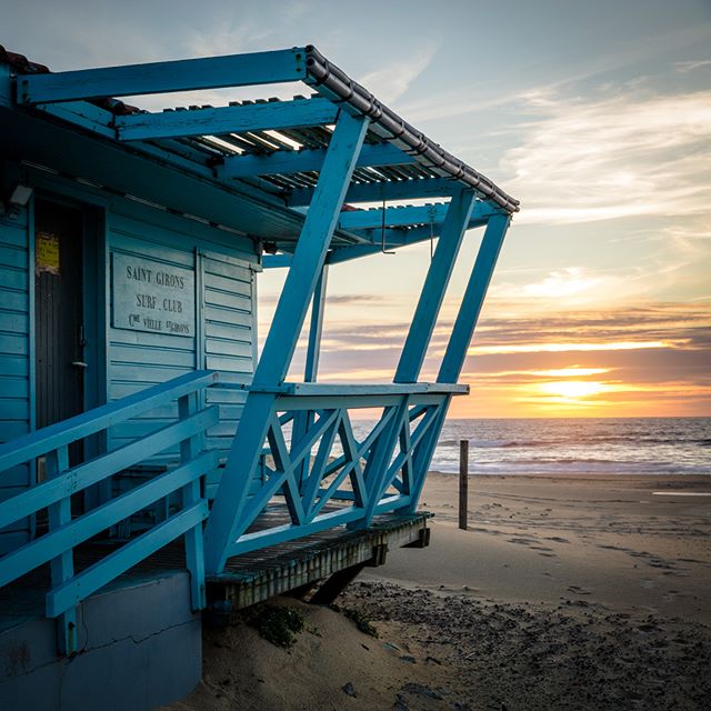 A blue surf club hut sits on a sandy beach at sunset, creating a peaceful coastal scene.
