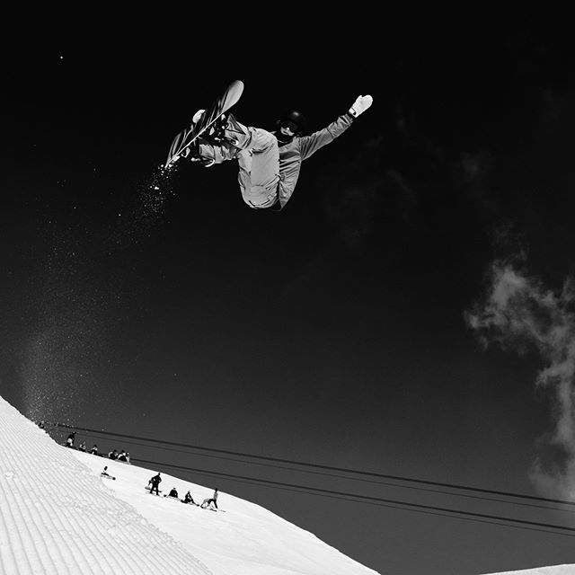 A snowboarder performs a trick mid-air against a snowy mountain backdrop in black and white.