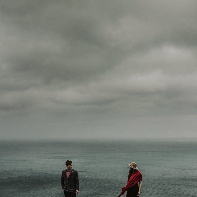 A couple stands on a cliff overlooking the ocean on an overcast day, creating a moody and romantic scene.