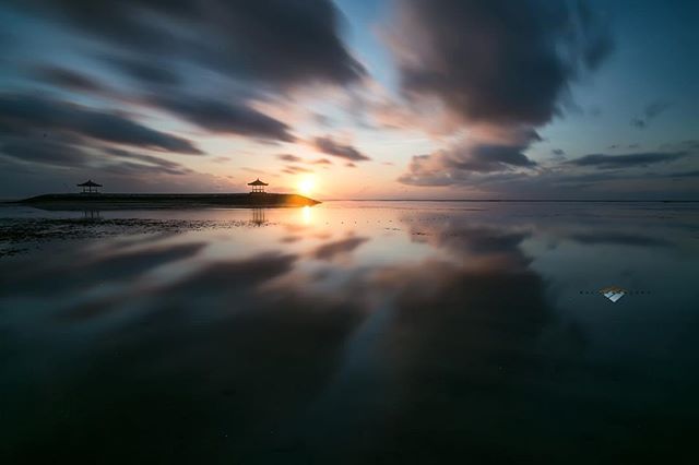 A serene sunset over the ocean with a reflective surface and a gazebo silhouetted against the setting sun.