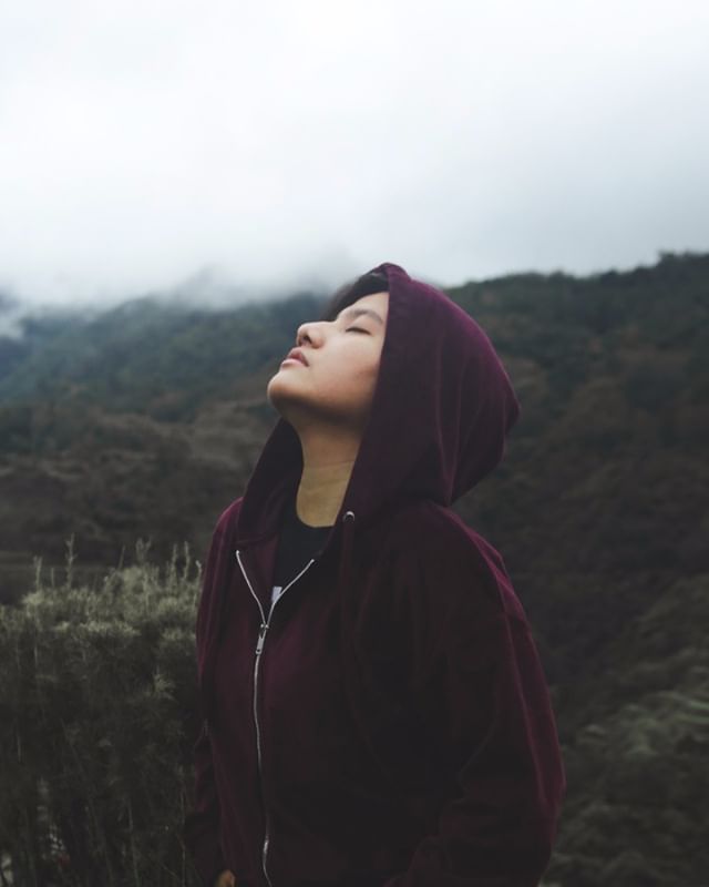 Woman in a hoodie relaxes outside in nature with her eyes closed, enjoying the fresh air and tranquil scenery.