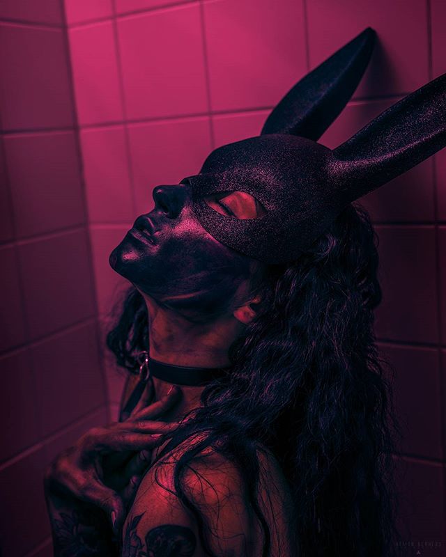 A woman wearing a bunny mask poses dramatically against a pink tiled wall in a moody, sensual artistic shot.