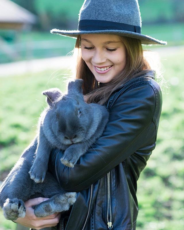 A smiling woman in a hat and leather jacket lovingly holds a gray rabbit in a sunny outdoor setting.