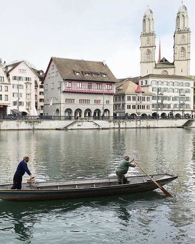 Two men row a boat on a river in Zurich, Switzerland, with the Grossmünster Church in the background.