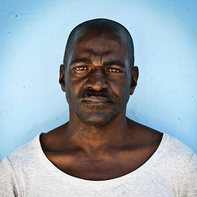 A serious dark-skinned man with a mustache gazes directly at the camera against a light blue wall.
