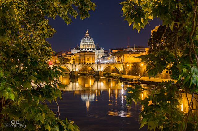 Illuminated St. Peter's Basilica and bridge over the Tiber River at night in Rome, framed by foliage.