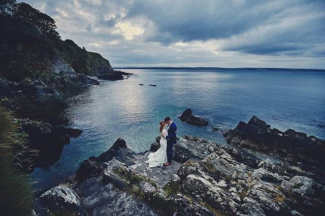 A bride and groom embrace on a rocky cliff overlooking the ocean on their wedding day.